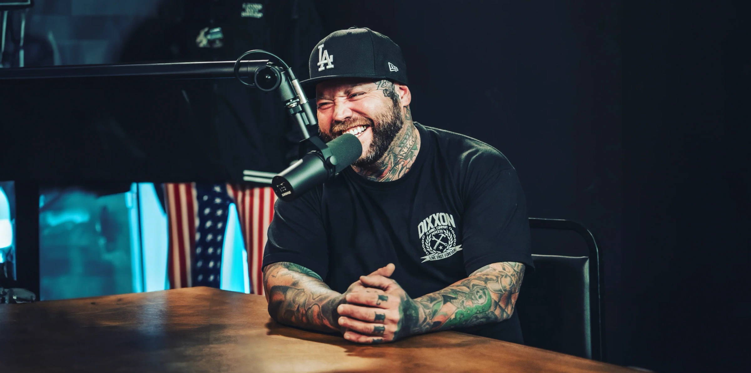 A tattooed man in a Dixxon Flannel Company tee and a black cap laughing into a studio microphone, from the Our Story page.