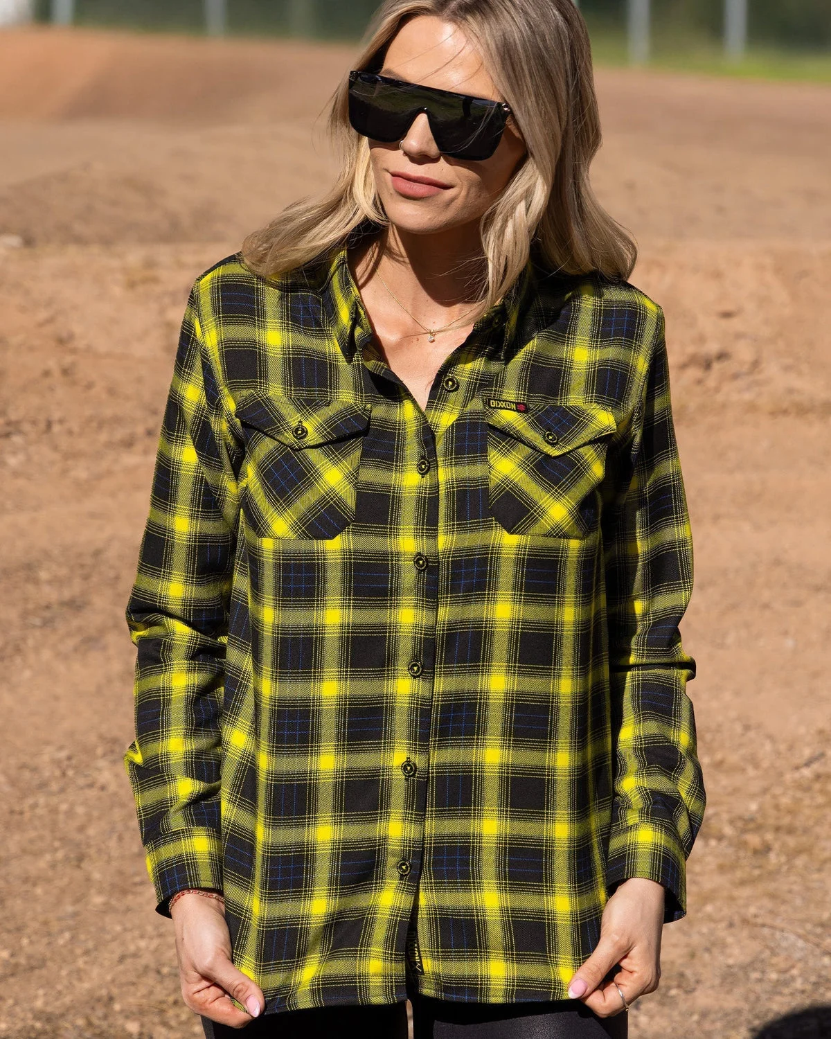 A woman in sunglasses wearing a black and yellow plaid Dixxon flannel on a dirt track.